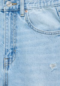 Jean en denim bleu clair en gros plan montrant la poche avant, le bouton métallique, les coutures et une petite déchirure usée sur le tissu.