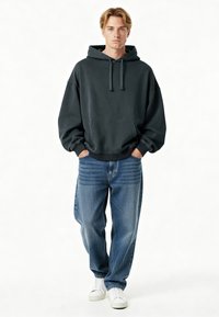 Sötét szürke, túlméretezett kapucnis pulóver, kenguru zsebbel és húzózsinórral, világoskék, bő nadrággal és fehér tornacipővel párosítva.