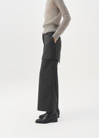 Črne široke hlače z raztrganimi robovi, v kombinaciji s svetlo sivo puhasto jopico. Model stoji vstran, z rokami v žepih, obut v črne klinaste čevlje.