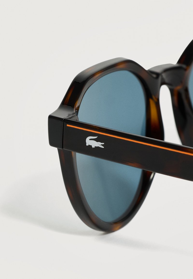 Sonnenbrille mit Schildpatt-Muster, blauen Gläsern und schwarzem Bügel mit orangem Streifen und weißem Krokodillogo.