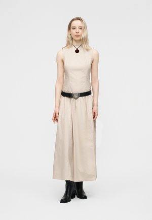 Femme portant une robe beige sans manches avec une ceinture noire à boucle en forme de cœur, des bottines noires et un collier à rose noire, debout sur un fond blanc.