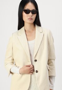 Beige corduroy-takk som har en notch-krage, to svarte knapper og avslappede ermer. Stylet med en lys genser under og solbriller.