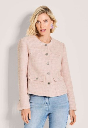 Blonde vrouw die een lichtroze gestructureerd jasje met knopen en voorzakken draagt, gecombineerd met een lichtblauwe jeans, staand tegen een beige achtergrond.