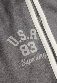 Grå sweatshirt med en hvid stribet lynlås, med trykt tekst "U.S.A 83" og "Superdry" i en kontrasterende lys farve. Blød stoftekstur.