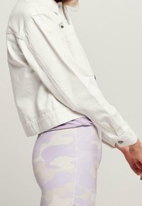 Bílá denim bunda s dlouhými rukávy, knoflíkovými detaily a boční kapsou. V kombinaci s legínami v lilákovém maskáčovém vzoru. Viditelné jemné textury látek.