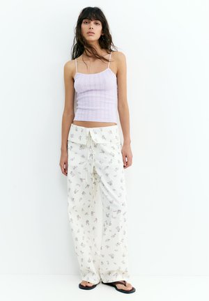 Jeune femme aux longs cheveux foncés portant un débardeur côtelé violet clair, un pantalon blanc à larges jambes à fleurs, et des tongs noires, debout contre un mur blanc.