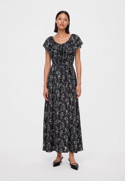 Woman stands wearing a black floral maxi dress with ruffled short sleeves and black pointed-toe slingback shoes on a white background.