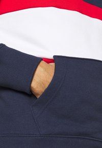Main dans la poche d'un sweat-shirt bleu marine, sur un fond de chemise à blocs de couleur rouge, blanc et bleu marine.