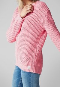 Różowy sweter z dzianiny o rypsowym wzorze, z dekoltem w kształcie V i bocznymi rozcięciami. W połączeniu z jasnoniebieskimi dżinsami i białym detalem metki.