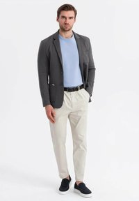 Hombre vestido con blazer gris, suéter azul claro, pantalones beiges y zapatos negros sin cordones, de pie con una mano en el bolsillo, sobre un fondo blanco.