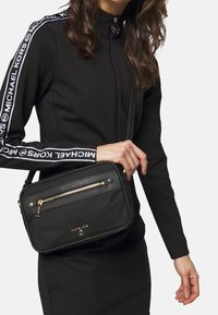 Schwarze Crossbody-Tasche aus glattem Stoff mit goldigem Reißverschluss, Fronttasche und geprägt Logo. Verstellbarer Tragegurt und kastenförmiges Design.