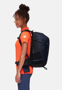 Schwarzer und marineblauer Rucksack mit mehreren Riemen, einem strukturierten Design und einem vorderen Reißverschlussfach, getragen von einer Person in einem leuchtend orangefarbenen Shirt.