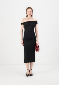 Svart off-shoulder kjole med rynk, tettsittende silhuett, kne-lang, brukt med svarte ankelrem sandaler og en liten rød veske.