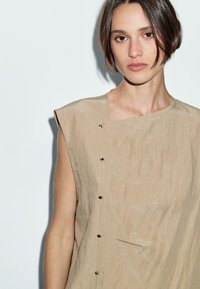 Beige Leinenoberteil mit rundem Ausschnitt, asymmetrischem Design, ärmellosem Schnitt und dekorativen Metallknöpfen an der Vorderseite.