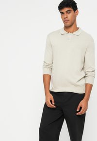 Camisa polo de manga longa bege com colarinho, apresentando uma pala de três botões e punhos canelados, combinada com calças pretas. Textura suave.