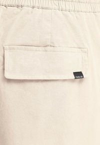 Beige shorts feature an elastic waistband and a back pocket with a black label. The fabric is smooth with a subtle texture.