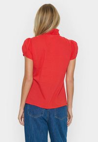 Blusa rossa con colletto arricciato, maniche a sbuffo corte e una texture liscia; abbinata a jeans a gamba larga blu con tasche posteriori.