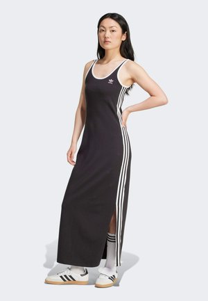 Robe maxi sans manches noire avec des accents blancs et trois rayures blanches sur les côtés, dotée d'une fente sur le côté. Portée avec des chaussures de sport blanches.