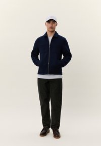 Ung mann iført en marineblå glidelåsjakke, hvit skjorte, svarte bukser, mørkebrune sko og en lys grå baseballcap, stående med hendene i lommene.