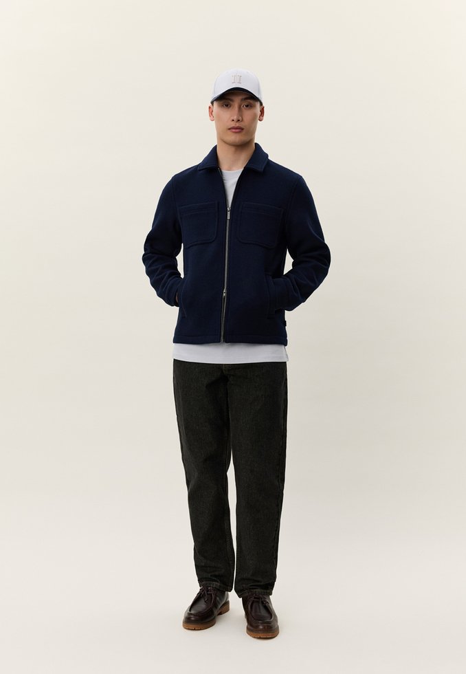 Ung mann iført en marineblå glidelåsjakke, hvit skjorte, svarte bukser, mørkebrune sko og en lys grå baseballcap, stående med hendene i lommene.