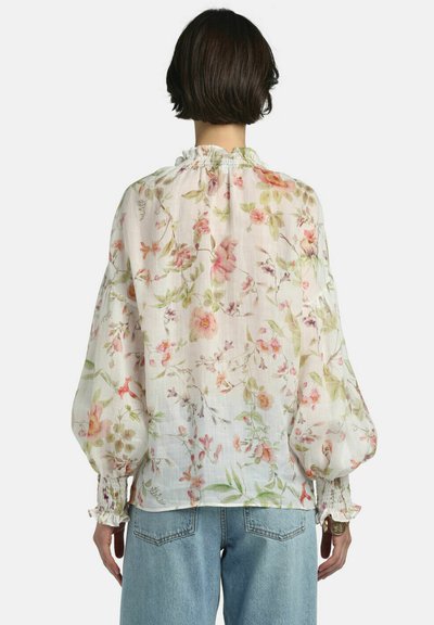 Kvinde med kort mørkt hår iført en gennemsigtig hvid bluse med pink og grønne blomsterprint og puffede ærmer, kombineret med lyseblå jeans.