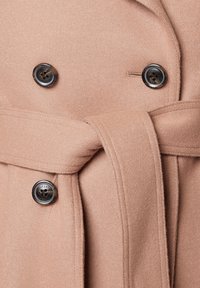 Różowopomarańczowy wełniany płaszcz z kołnierzem, podwójnym zapięciem na dwie ciemne guziki oraz paskiem z materiału zawiązanym w talii.