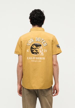 Mężczyzna z kręconymi włosami noszący musztardowo-żółtą koszulkę z krótkim rękawem z napisem "Von Dutch California Motorcycle Club" i grafiką czaszki w hełmie w płomieniach na plecach.