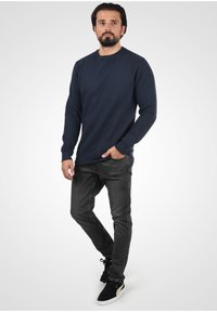 Sudadera texturizada azul marino con detalle acanalado, cuello redondo y mangas largas, combinada con jeans gris ajustados y zapatillas negras.