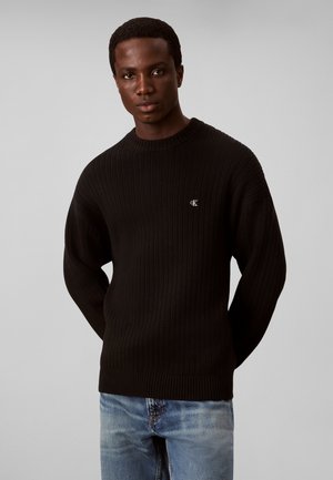 Homme portant un pull côtelé noir avec un petit logo blanc "cK" sur la poitrine et un jean bleu clair, debout devant un fond gris uni.