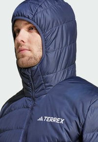 Marineblå, puffy hættejakke med høj krave, quiltet tekstur og broderet "TERREX" logo på brystet, der fremhæver en tæt pasform.