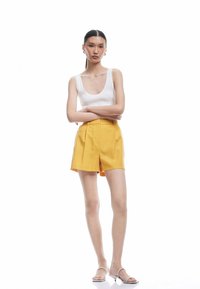 Pantaloni corti gialli su misura con tasche laterali, frontale piatto e tessuto liscio. Abbinati a una canotta bianca a costine e sandali di colore chiaro.
