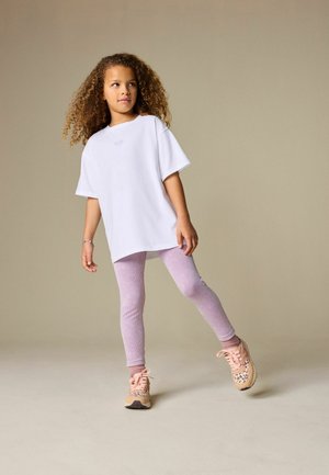 Lokkidega laps, kes kannab üle suuruses valget t-särki, helelilla retuuse, roosasid sokke ja beeže leopardimustriga kingi, seistes ühel jalal.