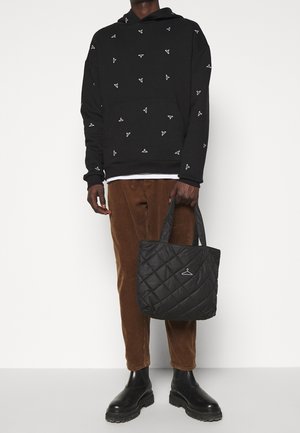 Sweatshirt noir avec des motifs blancs, pantalons en velours côtelé brun et un sac fourre-tout noir matelassé, avec un petit logo suspendu et des poignées solides.