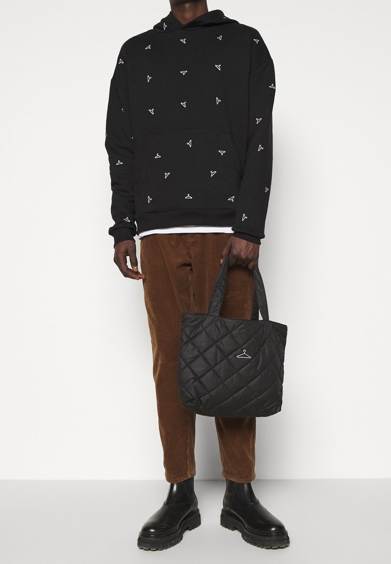 Sweatshirt noir avec des motifs blancs, pantalons en velours côtelé brun et un sac fourre-tout noir matelassé, avec un petit logo suspendu et des poignées solides.