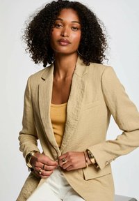Blazer di lino beige con una vestibilità rilassata, tasca frontale singola e bottoni. Abbinato a un top giallo e dettagli di gioielli dorati.