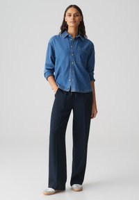 Camicia in denim di blu medio con colletto e taschino sul petto. Abbinata a pantaloni tapered blu scuro e sneakers bianche.