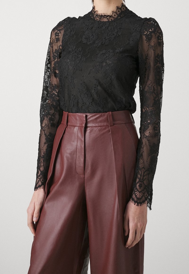 Blouse noire en dentelle à manches longues avec motifs floraux, associée à un pantalon en cuir bordeaux, taille haute et jambes larges.