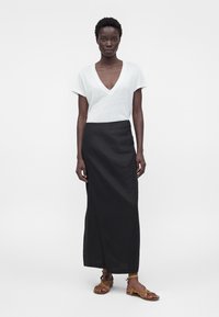 Ženska stoji in nosi belo majico z V-izrezom in kratkimi rokavi, črno krilo do gležnjev z bočnim razporkom in rjave ravne sandale z trakci.