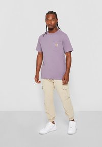Lavender short-sleeve T-shirt with a chest pocket, paired with beige cargo pants and white sneakers. Smooth cotton texture, casual fit.