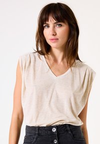 Helles beige, ärmelloses V-Ausschnitt-Oberteil mit subtiler Textur und leichten Schulterfalten. Kombiniert mit schwarzen, hochtaillierten Jeans und sichtbaren Knöpfen.