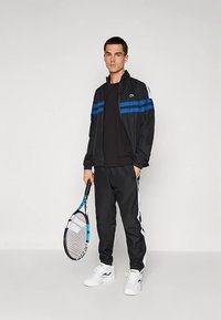 Μαύρη αθλητική φόρμα με μπλε ρίγες, ζακέτα με φερμουάρ και αντίστοιχο παντελόνι. Κρατάει μια ρακέτα τένις, διαθέτει λευκές λεπτομέρειες και αθλητικά παπούτσια.