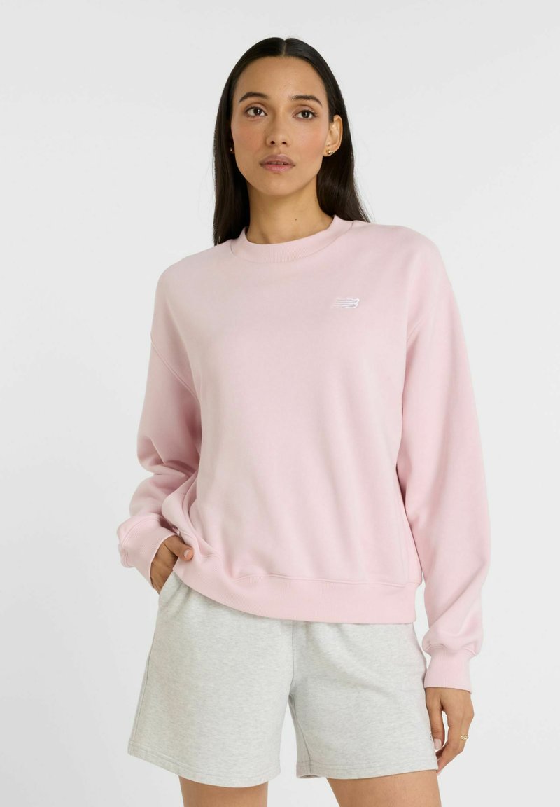 Sudadera rosa de corte relajado, con puños acanalados y cuello redondo; presenta un pequeño logo blanco en el pecho y una textura suave.