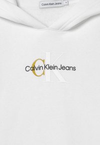 Sudadera blanca con el logo "Calvin Klein Jeans" en negro sobre grandes letras blancas y doradas "CK" en la parte frontal, etiqueta de tamaño visible en el interior del cuello.