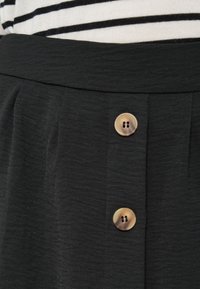 Μαύρη φούστα από ύφασμα με υφή, με λείο ζωνάρι στη μέση και δύο καφέ κουμπιά ως κύρια διακοσμητικά στοιχεία.