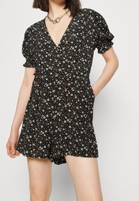 Femme portant une combinaison courte noire à motifs floraux, à manches courtes, avec un décolleté en V profond et un collier en chaîne, la main dans la poche, sur un fond uni.