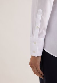 Hånd hvilende ved siden af en hvid skjortemanschet med knapper og to knapper på manschetpladen, mod en neutral baggrund.