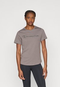 Gray cotton t-shirt with rounded neckline and short sleeves, featuring an embroidered "Champion" logo across the chest. Paired with black leggings.