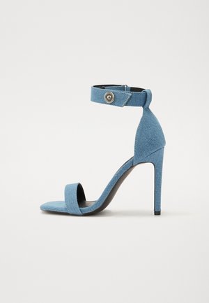 Modré džínsové sandále na vysokom opätku s remienkom okolo členka a zapínaním na gombík. Majú štvorcový špičku a čiernu podrážku pre kontrast.