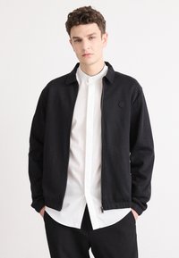 Schwarze Zip-Jacke mit Kragen, mit einem Logo auf der Brust, kombiniert mit einem weißen Hemd und schwarzen Hosen. Glatter Stoff, lässiges Design.