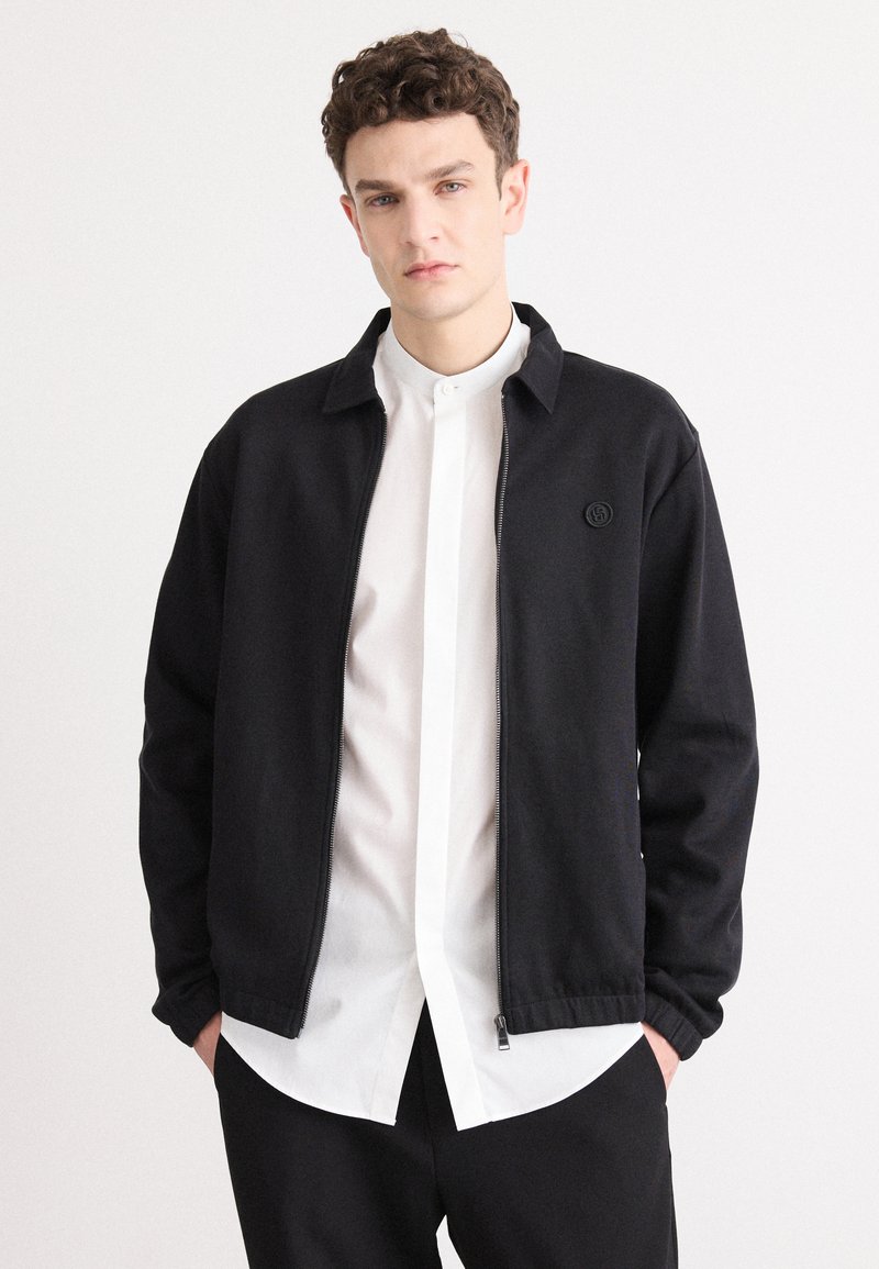 Schwarze Zip-Jacke mit Kragen, mit einem Logo auf der Brust, kombiniert mit einem weißen Hemd und schwarzen Hosen. Glatter Stoff, lässiges Design.